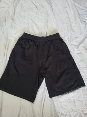 Svarta träningsshorts från SOC - Svarta träningsshorts från SOC med elastisk midja och avslappnad passform. Perfekta för gymmet eller löprundan. Lätta och bekväma med diskret logga på benet. Klassisk design utan extra detaljer.