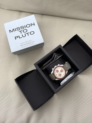Swatch x Omega Mission to Pluto klocka - Säljer en Swatch x Omega Mission to Pluto från Moonswatch-kollektionen. Klockan har en vit boett i biokeramik, beige urtavla med röda detaljer och grått textilband. Kommer i originalbox med tillbehör. Perfekt för dig som gillar unika accessoarer.