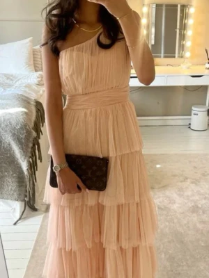 Ljusrosa oneshoulder långklänning - Romantisk ljusrosa långklänning med volanger och oneshoulder-design. Klänningen har flera lager av tunt tyg som ger en drömmig känsla och markerad midja. Perfekt för speciella tillfällen där du vill sticka ut.