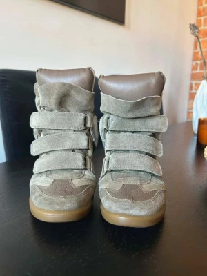 Beige höga sneakers med kardborre - Unika höga sneakers i beige mocka och brunt läder med flera breda kardborreband. Snygg design med vadderad plös och gummisula i ljusbrunt. Perfekta för dig som vill sticka ut med en cool och trendig look. Storlek 38 äkta isabel marants 