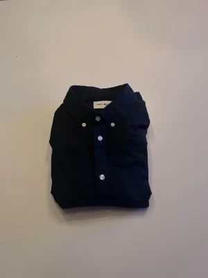 Mörkblå skjorta från East West - Klassisk mörkblå skjorta från East West med vita knappar och button down-krage. Skjortan har lång ärm och är tillverkad i mjuk bomull, perfekt för en clean och stilren look.