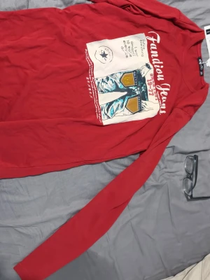 Röd långärmad topp Fandian Jeans - Cool röd långärmad topp från Fandian Jeans med tryck framtill som visar ett par jeans och text. Rund halsringning och mjukt material som känns skönt mot huden. Perfekt för dig som gillar streetwear och vill sticka ut lite extra.