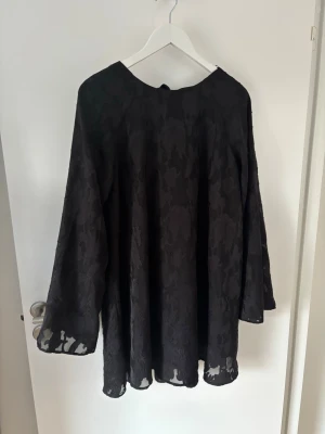Svart blommig långärmad klänning - Svart klänning med lång ärm och diskret blommigt mönster i tyget. Klänningen har en lös passform och är tillverkad i ett lätt, genombrutet material som ger en elegant känsla. Perfekt för dig som gillar stilrena och unika detaljer.