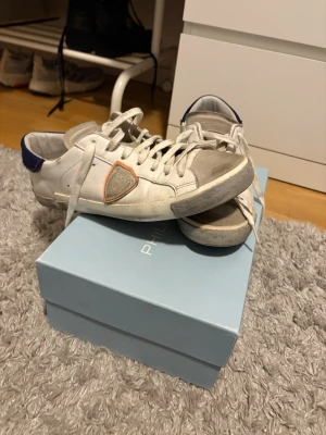 Philippe Model vita sneakers - Snygga vita sneakers från Philippe Model med grå detaljer på tån och en blå hälflik där märket står broderat. Skorna har en ikonisk sköldlogga på sidan och vita skosnören. Följs med kartong och extra snören. Priset inte hugget i sten va inte rädda att höra av er. Mvh 