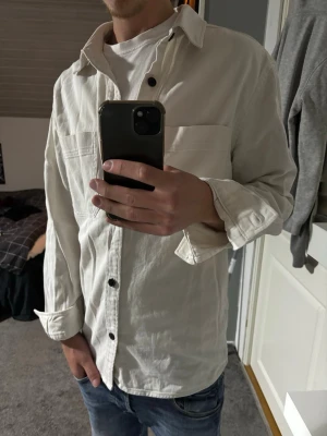 Vit Overshirt  - Stilren beige overshirt med svarta knappar och bröstficka. Skjortan har lång ärm och klassisk krage, perfekt att bära öppen över en t-shirt. Tillverkad i ett mjukt bomullstyg som ger en avslappnad look.