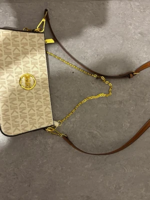 Beige axelväska från Michael Kors - Snygg beige axelväska från Michael Kors med MK-logga i guld framtill. Väskan har ett mönstrat tyg med bruna detaljer, guldfärgade kedjor och justerbar axelrem i brunt läder. Perfekt för dig som gillar stilrena accessoarer med lyxig känsla.