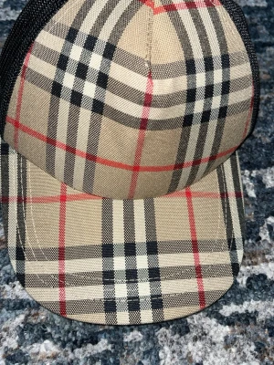 Burberry keps - Snygg keps i klassiskt beige, svart och rött rutmönster med svart mesh på baksidan. Kepsen har böjd skärm och justerbar rem bak. Perfekt för dig som gillar streetwear och vill sticka ut med en ikonisk look.