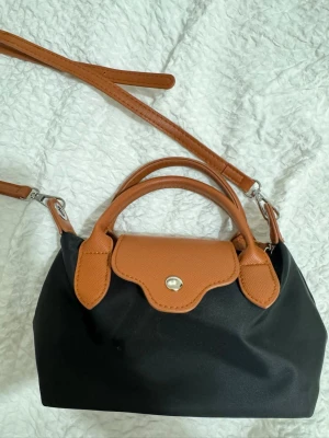 Svart och brun handväska Longchamp - Snygg svart handväska från Longchamp med bruna läderdetaljer och avtagbar axelrem. Väskan har klassisk form, silverfärgad knapp och dubbla handtag. Tillverkad i slitstark nylon och läder, perfekt för dig som gillar stilrena accessoarer.