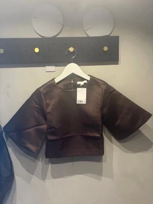 Brun croppad topp i satin - Snygg brun croppad topp med vida korta ärmar och rund halsringning. Tillverkad i glansigt satinmaterial som ger en lyxig känsla. Perfekt att styla med höga jeans eller kjol för en trendig look.