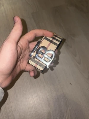 Beige/svart bälte - Elegant bälte i klassiskt rutmönster med beige bas och svarta, vita samt röda ränder. Bältet har ett silverfärgat metallspänne format som dubbla bokstäver “BB”, vilket ger en stilren och exklusiv look. Insidan är svart med präglat hästmotiv. Tillverkat i ett läderliknande material med justerbar längd för bekväm passform.
