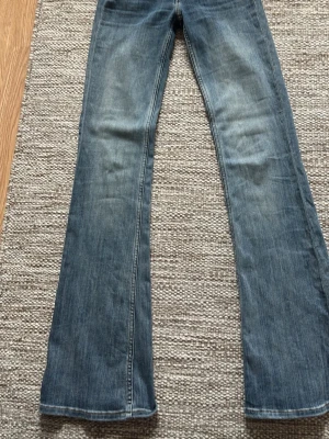 Blå bootcut jeans med slitningar - Säljer ett par blå bootcut jeans med lätt slitna detaljer och klassisk femficksmodell. Jeansen har normalhög midja, bälteshällor och dragkedja med knapp. Materialet är denim i bomull med lite stretch för skön passform.