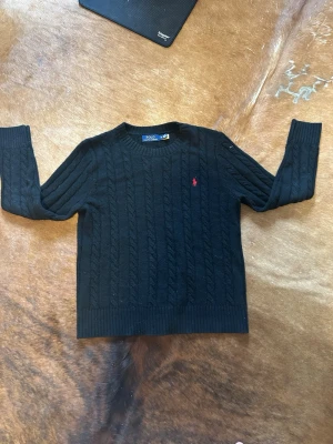 Mörkblå kabelstickad tröja Polo Ralph Lauren - Kabelstickad mörkblå tröja från Polo Ralph Lauren med rund halsringning och ribbade muddar. Klassisk röd broderad logga på bröstet. Tillverkad i mjukt material som passar perfekt till lager på lager. Snygg och tidlös design. 