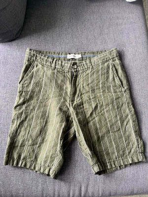  linneshorts - Snygga gröna shorts från Dressmann i linne med vita ränder. Klassisk passform med fickor fram och bak samt bälteshällor. Perfekta för varma dagar och en avslappnad stil.