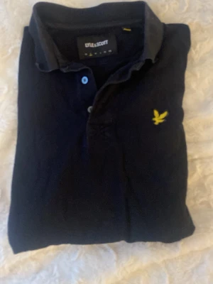 Svart piké från Lyle & Scott S - Klassisk svart pikétröja från Lyle & Scott med gul broderad logga på bröstet. Skjortan har krage, knappar framtill och är tillverkad i mjuk bomull. Perfekt för en clean och stilren look.
