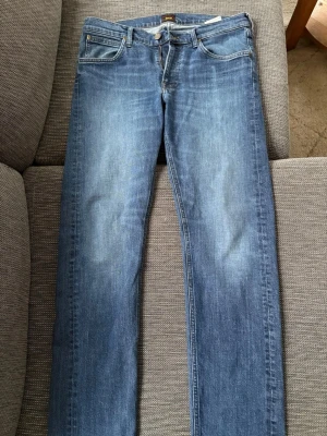 Blå raka Lee jeans  - Säljer ett par blå jeans med rak passform och klassisk tvätt. Jeansen har fem fickor, normal midja och subtila slitningar framtill för en avslappnad look. Perfekta till vardagsstilen och tillverkade i mjukt denimtyg.