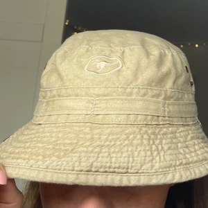Bucket hat  - Cool beige bucket hat i storlek S med en broderad känguru fram och knappar som går att knäppa upp kanten med på sidan. Hatten är i använt skick, därav det låga priset (40kr + frakt eller så kan jag mötas i Stockholm)!