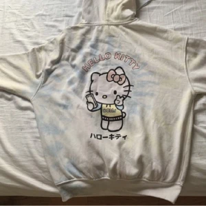 Hello Kitty huvtröja - Hello Kitty, bra kvalitet, hoodie, grundfärgen är beige och så finns det lite blå, gul, vitt, rosa. Det finns Hello Kitty och japanska text på baksidan av tröjan.
