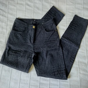 Svarta/mörkgrå högmidjade mjuka mönstrade byxor - snakeskin - storlek 36 - Snygga svarta/mörkgrå högmidjade byxor med guldfärgade detaljer från H&M. Sytt snakeskininspirerat mönster. Tyget är en aning nopprigt/ludet på vissa ställen. 55% viskos 41% bomull 4% elastan. Storlek 36. 