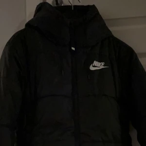 Nike - svart vadderad jacka med huva och Nike logga - Hej jag säljer min jätte snygga Nike jacka. Den köptes i slutet av vintern/våras så den  användes bara någon gång eftersom det blev sommar så den är i nyskick. Passar perfekt till vintern för den håller en varm. Skriv för frågor om betalning osv!