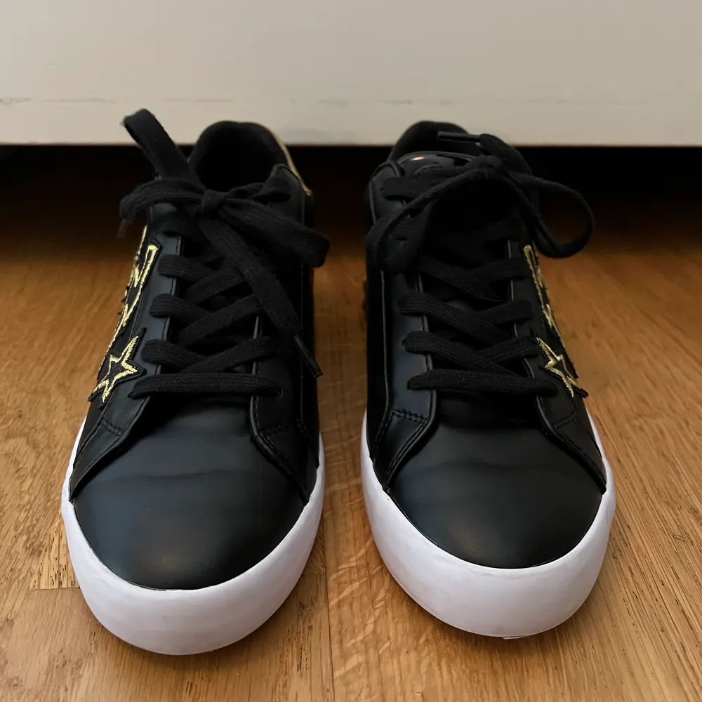 Säljer mina jätte coola Guess sneakers i svart med  guld detaljer, säljer pga att de är för små. Sneakersarna är använda ett fåtal gånger och är i nyskick. På baksidan står Guess i guld och det är en stjärna, blixt och ett hjärta i guld på ena sidan på båda skorna. Skorna är i storlek 39.. Skor.