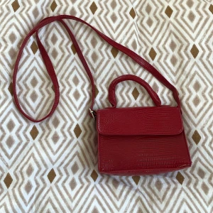 Röd väska i skinnimitation - Fin mörkröd väska i skinnimitation med justerbart band samt handtag. I nyskick. 19 cm x 15 cm x 9 cm. Säljes då jag har för många väskor.