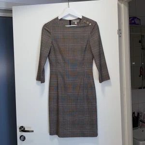 Grårutig klänning från H&M strl 34 - Väldigt söt grårutig klänning från H&M i strl 34/XS. Klänning är använd men väldigt sparsamt så nästan som ny. Midjan är lite smalare vilket ger en väldigt smickrande form. Har även (som går att ses på sista bilden) guldiga knappar på båda ärmarna och vid högra axeln. 