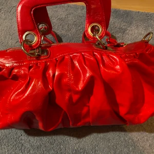 Röd Puccini Väska - Säljer den här jättefina röda väskan då den inte kommer till användning. Superfint skick och kommer även med en matchande väsk rem. Jag skulle säga att den är ganska stor och får plats med mycket saker. Metall detaljerna är silvriga.