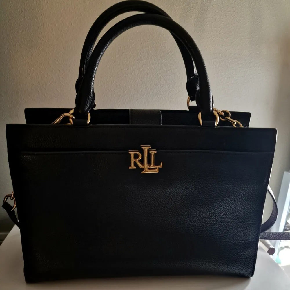 Klassisk svart handväska från Lauren Ralph Lauren i skinn med detaljer i guld. Perfekt vid alla tillfällen - så väl som till fest som till vardag. Den är i nyskick då den använts vid enstaka tillfällen så det är ett rikligt kap!. Väskor.