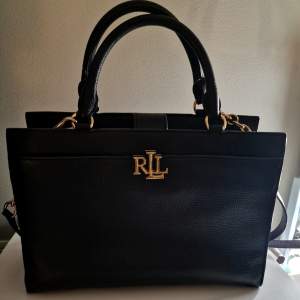 Klassisk svart handväska från Lauren Ralph Lauren i skinn med detaljer i guld. Perfekt vid alla tillfällen - så väl som till fest som till vardag. Den är i nyskick då den använts vid enstaka tillfällen så det är ett rikligt kap!
