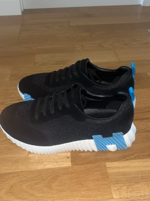 Svarta sneakers med blå detaljer - Snygga svarta sneakers med mesh- och mockaliknande material, vita räfflade sulor och blå detaljer på hälen och sidan. Skorna har klassisk snörning och låg profil som passar till många olika stilar. Perfekta för dig som gillar en modern och sportig look.