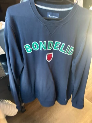 Mörkblå sweatshirt från Bondelid - Mörkblå sweatshirt från Bondelid med grön och vit text på bröstet samt ett rött och vitt sköldmärke. Klassisk rund halsringning och ribbade muddar vid ärmslut och nederkant. Perfekt för en avslappnad och stilren look.
