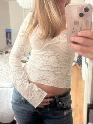 Vit spetstopp - Säljer en superfin vit omlottopp i spets med v-ringning och långa ärmar. Toppen är croppad och har ett romantiskt blommigt spetsmönster som ger en fräsch och trendig look. Perfekt att styla med jeans eller kjol för en snygg outfit. Använd en hån och är i perfekt skick!