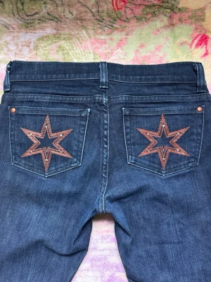 Lågmidjade Bootcut Victoria Beckham jeans - Snygga mörkblå lågmidjade jeans med utsvängda ben och unika stjärnbroderier i kopparfärg på bakfickorna!😻Även små stenar/diamanter för extra detaljer, samt ”dVb” loggan. Jeansen har klassisk femficksdesign och bälteshällor! Perfekta för dig som vill sticka ut med coola detaljer🤍Passar så bra till alla plagg och är så vackra att ha på sig🫶🏻Du kommer få komplimanger! Storlek W26/L35, passar XS. Mått cm: 36 midja, 18 grenhöjd, 82,5 innerben🩷De är stretchiga och i ett mjukt material. Inga defekter❤️