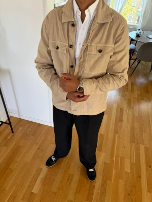 Beige overshirt med knappar från Gant  - Snygg beige overshirt i med klassisk krage och fyra framfickor. Stängs med svarta knappar framtill och har en avslappnad passform. Perfekt att bära över en skjorta för en stilren look. Är en large men sitter helt ok för M.