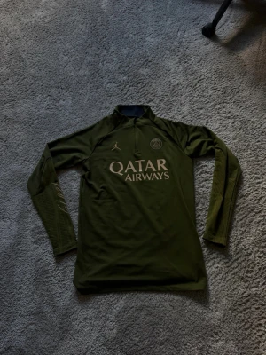 Mörkgrön PSG träningströja långärmad - Snygg mörkgrön långärmad träningströja från Paris Saint-Germain med halv dragkedja och tryckta loggor på bröstet. Tillverkad i ett lätt och funktionellt material som passar perfekt till träning eller fotboll. Stora Qatar Airways-trycket framtill.