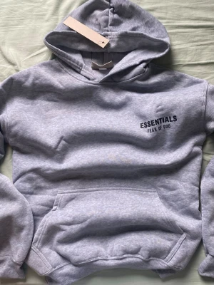 Grå Essentials Fear of God hoodie - Snygg grå hoodie från Essentials Fear of God med svart logga på bröstet. Klassisk känguruficka framtill och huva. Tillverkad i mjukt material som passar perfekt för en avslappnad streetwear-look.