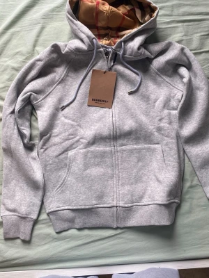 Grå zip hoodie från Burberry - Snygg grå zip hoodie från Burberry med klassiskt rutigt foder i huvan och dragsko. Hoodien har två stora fickor framtill och ribbade muddar vid ärmar och nederkant. Perfekt för en chill och stilren look.