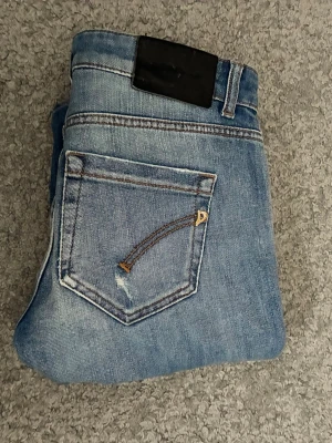Ljusblå jeans från Dondup med slitningar  - Säljer ett par ljusblå jeans med slitningar och hål på framsidan. Jeansen har klassisk femficksdesign, kontrastsömmar och en svart patch bak i midjan. Perfekta för en avslappnad och trendig look. Ny pris 3000