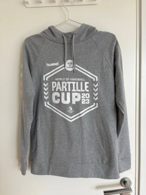 Grå Hummel Partille Cup hoodie - Snygg grå hoodie från Hummel med trycket 'Partille Cup 2023' och 'World of Handball' på bröstet. Klassisk huva med dragsko och långa ärmar. Perfekt för dig som gillar handboll och vill ha en sportig look.