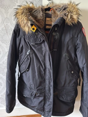 Svart parkasjacka med fuskpäls - Stilren svart parkasjacka från Parajumpers med avtagbar huva och tjock fuskpäls. Jackan har flera praktiska fickor, dragkedja och knappar framtill samt en gul detalj vid kragen. Perfekt för kalla dagar och har en patch på ärmen.