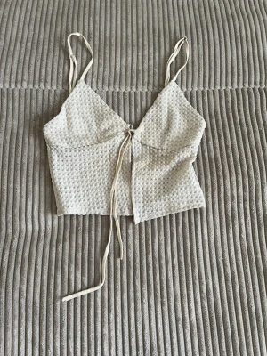 Beige croppad linnetopp med knyt - Trendig beige linnetopp med smala axelband och croppad passform. Toppen har ett diskret strukturerat mönster och knyt framtill som ger en snygg detalj. Perfekt för varma dagar och enkel att styla med jeans eller kjol.