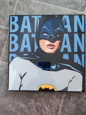 Batman tavla i pop art-stil - Snygg fyrkantig tavla med Batman-motiv i pop art-stil. Bakgrunden är blå med texten 'BATMAN' och motivet är tryckt på glas eller blank yta. Baksidan har upphängningsfäste och ramen är i trä. Perfekt för dig som gillar superhjältar och cool inredning.