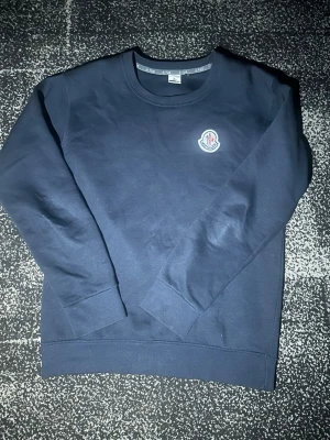 Mörkblå långärmad topp från Moncler - Säljer en stilren mörkblå långärmad topp från Moncler med rund halsringning och klassisk Moncler-logga på bröstet. Tillverkad i mjuk bomull som känns skön mot huden. Perfekt för dig som gillar enkel men snygg design.