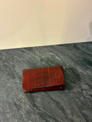 Brun plånbok i krokodilpräglat läder - Snygg begagnad brun plånbok i krokodilpräglat läder med flera kortfack, myntfack med dragkedja och ett fönster för ID-kort. Plånboken har tryckknappsstängning och en klassisk rektangulär form. Perfekt för dig som gillar stilrena accessoarer.