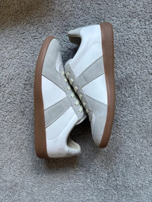 Vita och grå sneakers från Maison Margiela med äkta mocka och läder - Snygga sneakers i vitt och ljusgrått med klassisk design. Ovandelen är i en mix av slätt läder och mockadetaljer, och skorna har en brun gummisula. Perfekta för en clean och stilren look. Ljusbeiga skosnören och diskreta paneler ger en tidlös vibe.