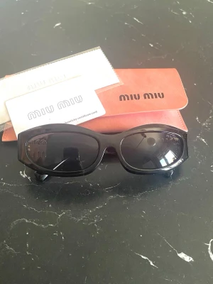 Svarta solglasögon från Miu Miu - Snygga svarta solglasögon från Miu Miu med rektangulär form och breda skalmar. På sidan sitter en cool gulddetalj med Miu Miu-loggan. Kommer med originalfodral och putsduk. Perfekta för dig som vill ha en trendig och exklusiv look.
