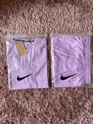Lila Nike träningsset - Säljer ett lila träningsset från Nike med matchande tröja och byxor. Setet har svarta Nike-loggor och är gjort i ett lätt och stretchigt material som passar perfekt för träning. Tröjan har rund hals och byxorna har resår i midjan.