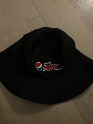 Svart bucket hat Pepsi Max - Cool svart bucket hat med broderad Pepsi Max-logga framtill och texten 'CHALLENGE YOUR TASTE' på baksidan. Hatten har bred skärm och är tillverkad i ett mjukt tygmaterial. Perfekt accessoar för dig som vill sticka ut med en unik och sportig stil.