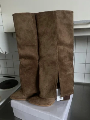 Bruna höga stövlar i mockaimitation - Helt nya snygga bruna stövlar med högt skaft och låg klack i trälook. Tillverkade i mjuk mockaimitation med slits på sidan för extra stil. Perfekta till jeans eller kjol för en trendig look. Dragfri design utan synliga detaljer framtill. Kan mötas i Södermalm eller tc. 