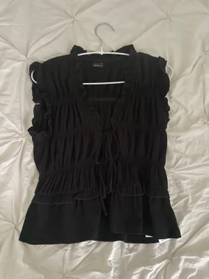 Svart blus med volang och knyt - Säljer en svart blus med volangdetaljer och knyt framtill. Blusen har v-ringning, ärmlös design och smockade partier som ger en snygg passform. Perfekt att styla med jeans eller kjol för en trendig look.